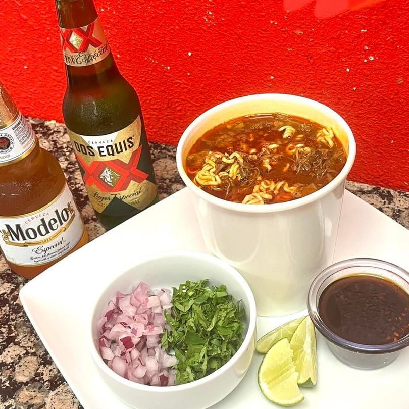 My Taco House birria ramen with chopped onions, cilantro, limes, and two Mexican beers.