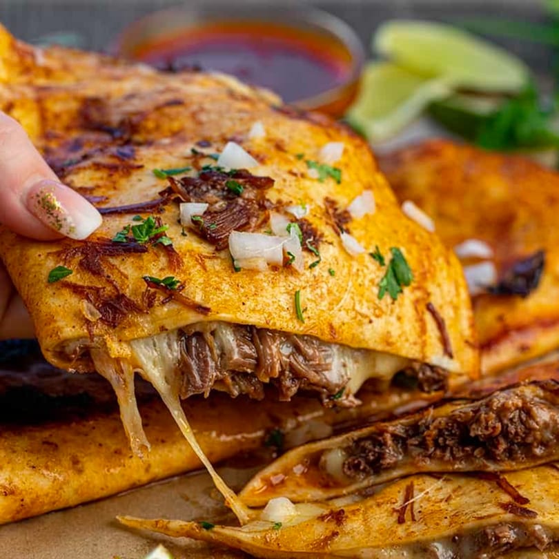 Hand holding a cheesy birria quesadilla from My Taco House, with dipping sauce and lime in background.