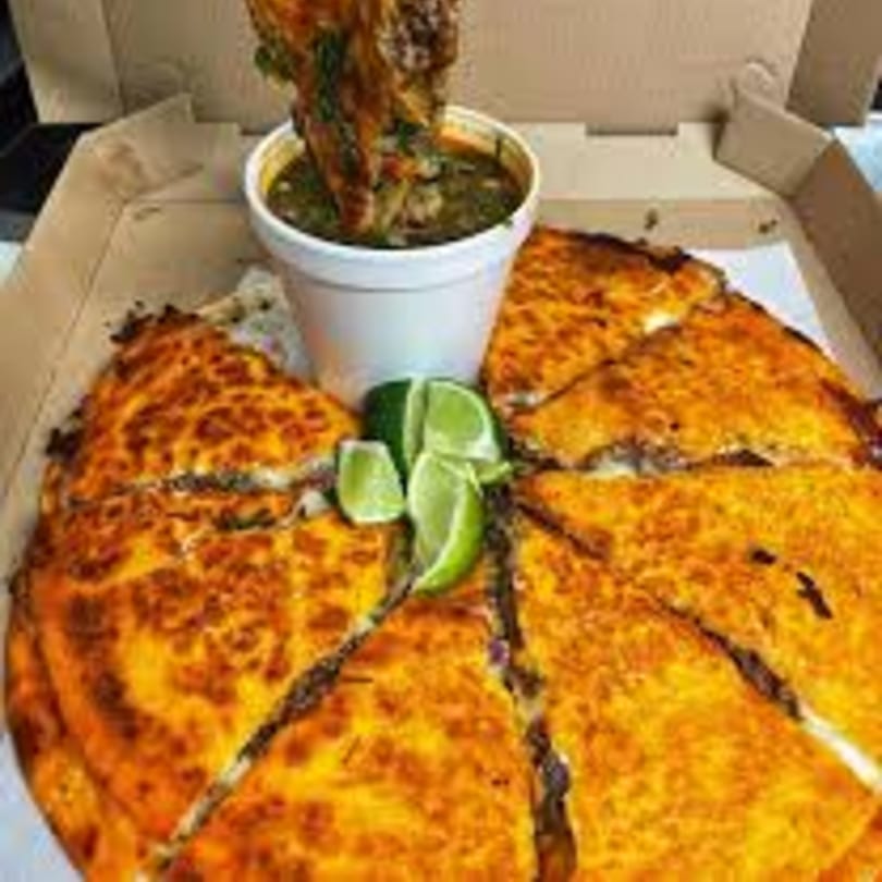 My Taco House birria pizza with a side of consommé and lime wedges in a takeout box.