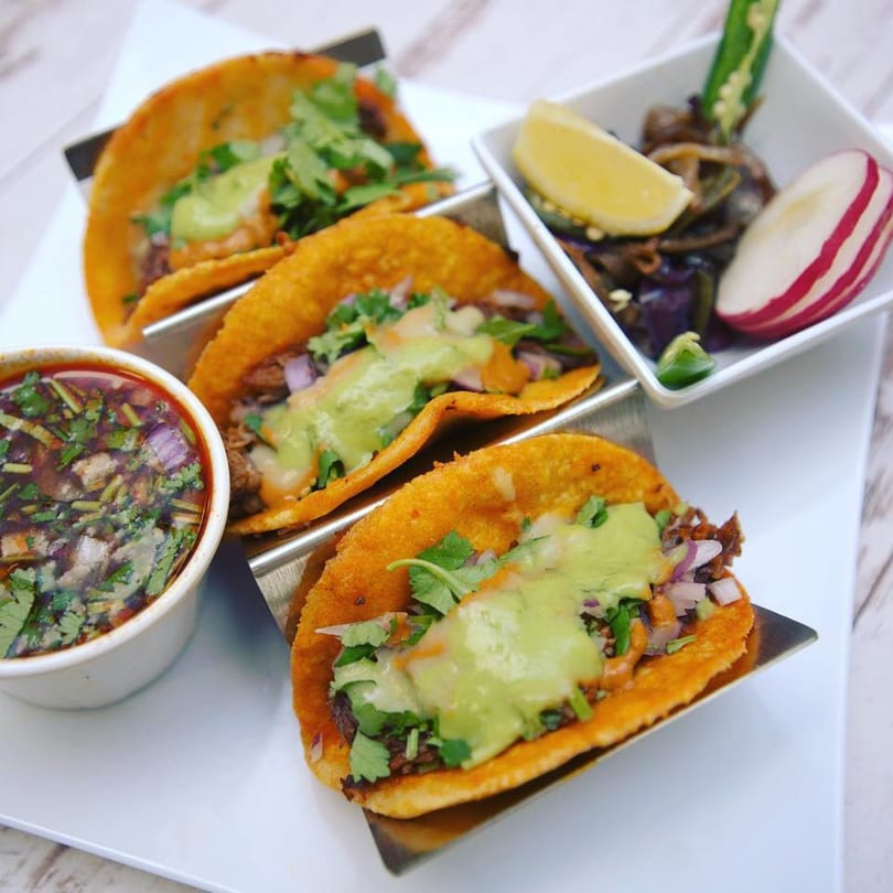 Three birria tacos with green sauce and cilantro, served with a side of consommé and pickled vegetables.